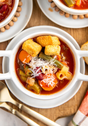 Up close image of minestrone soup in a white bowl with parmesan cheese and croutons on top.