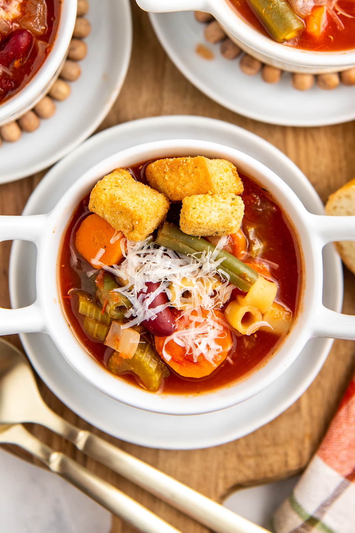 Up close image of minestrone soup in a white bowl with parmesan cheese and croutons on top.