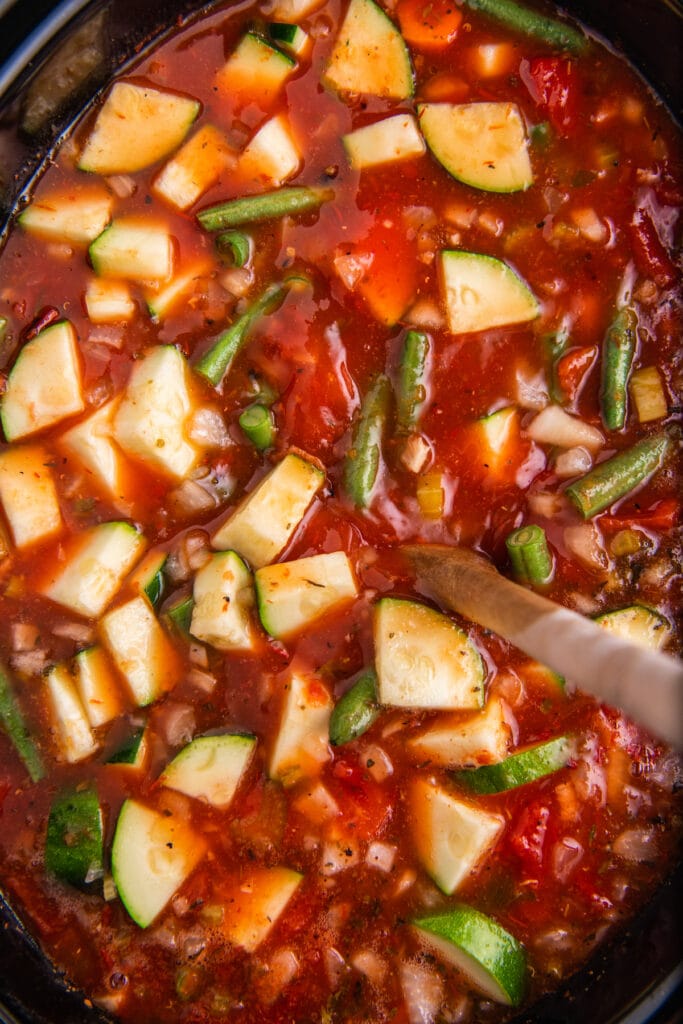 A brown wooden spoon stirring minestrone soup in a crockpot.