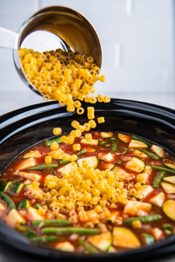 Pasta being poured into a crockpot filled with soup.