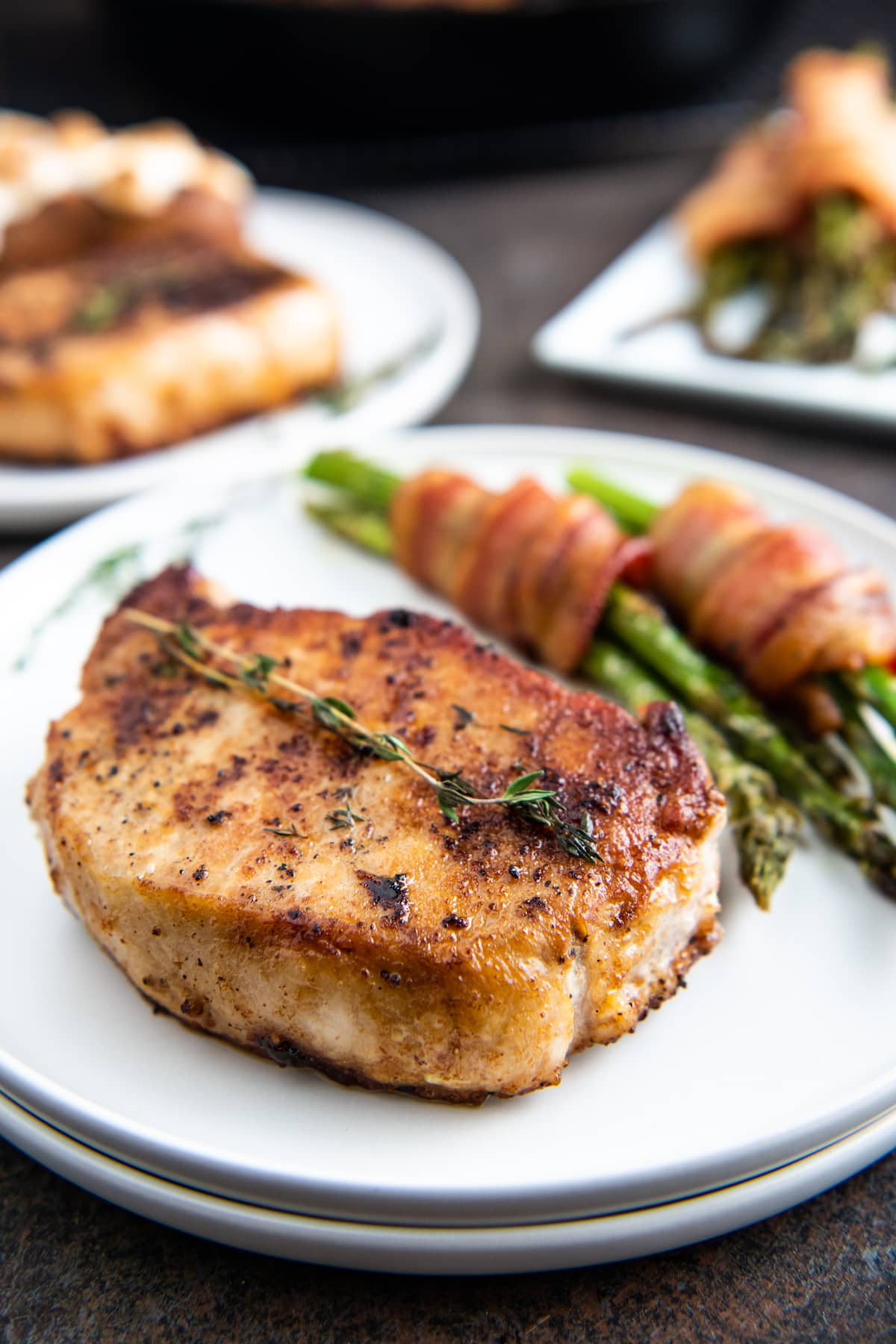 A seared pork chop on a white plate with asparagus wrapped with bacon.