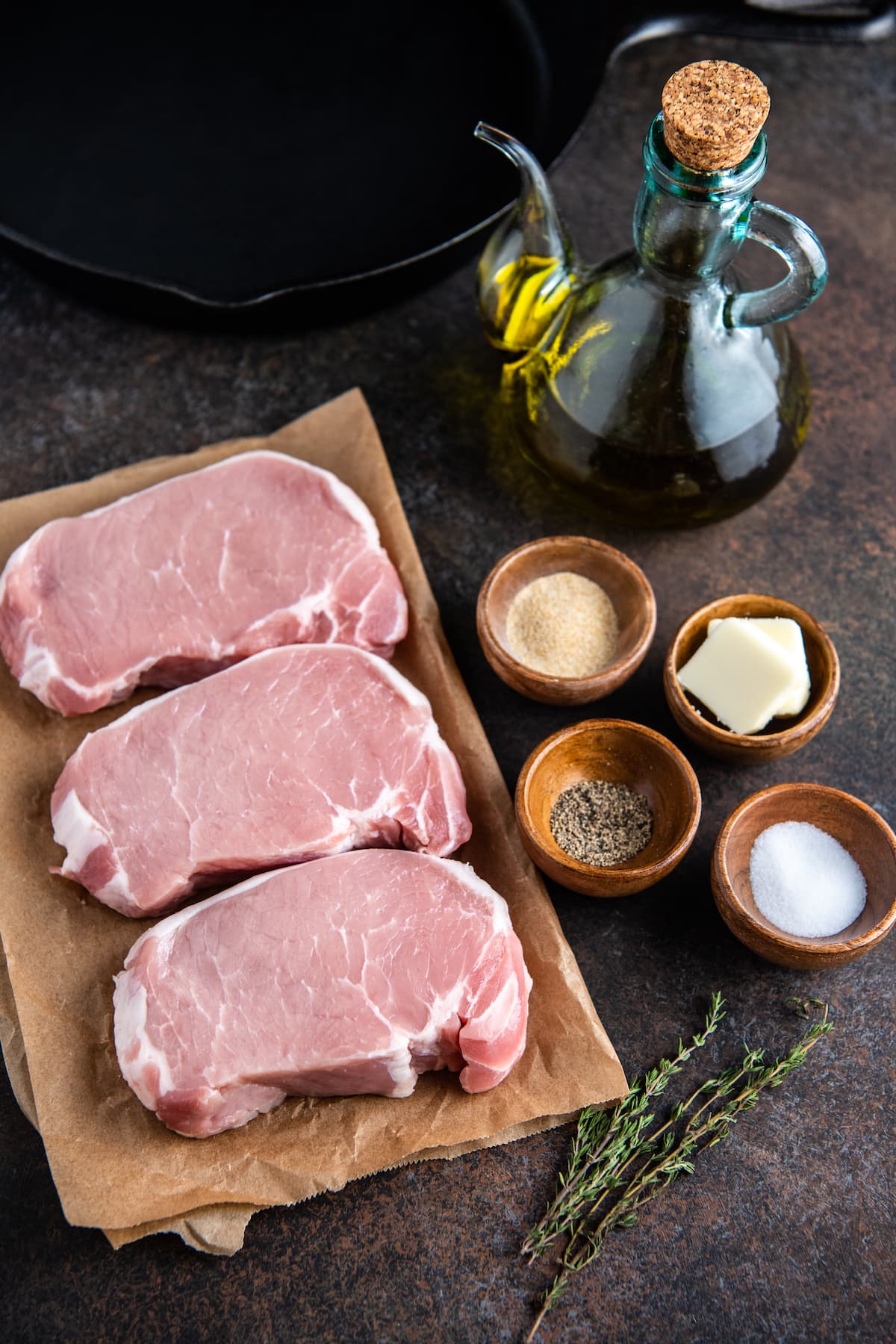 Three pork chops on brown parchment paper with seasonings in bowls and fresh thyme.