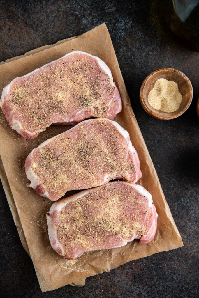 Three pork chops on a piece of parchment paper with seasonings on top.