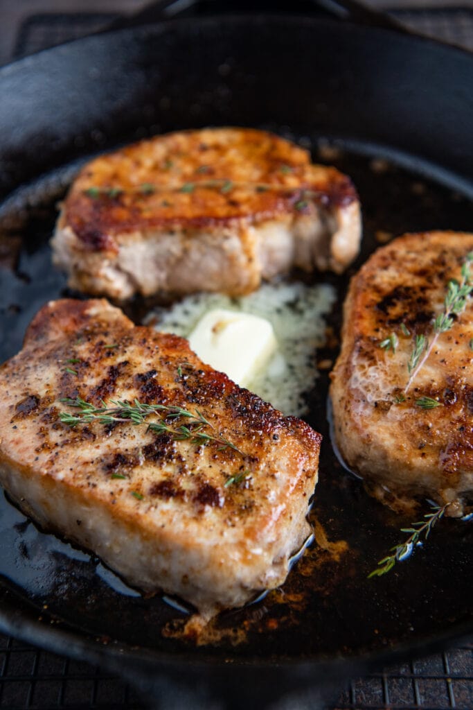 Three pork chops in a cast iron skillet with butter and herbs.