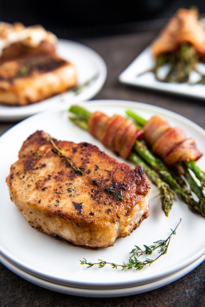 A pan fried pork chop on a white plate with thyme and bacon wrapped asparagus.