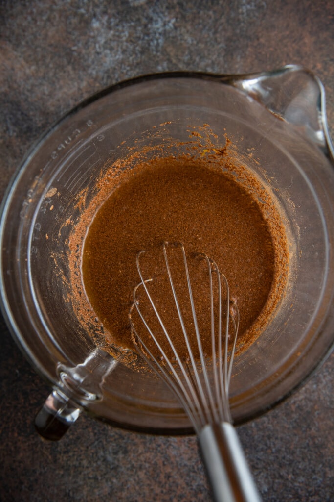 Seasonings, brown sugar, maple syrup and orange juice in a glass bowl with a whisk.