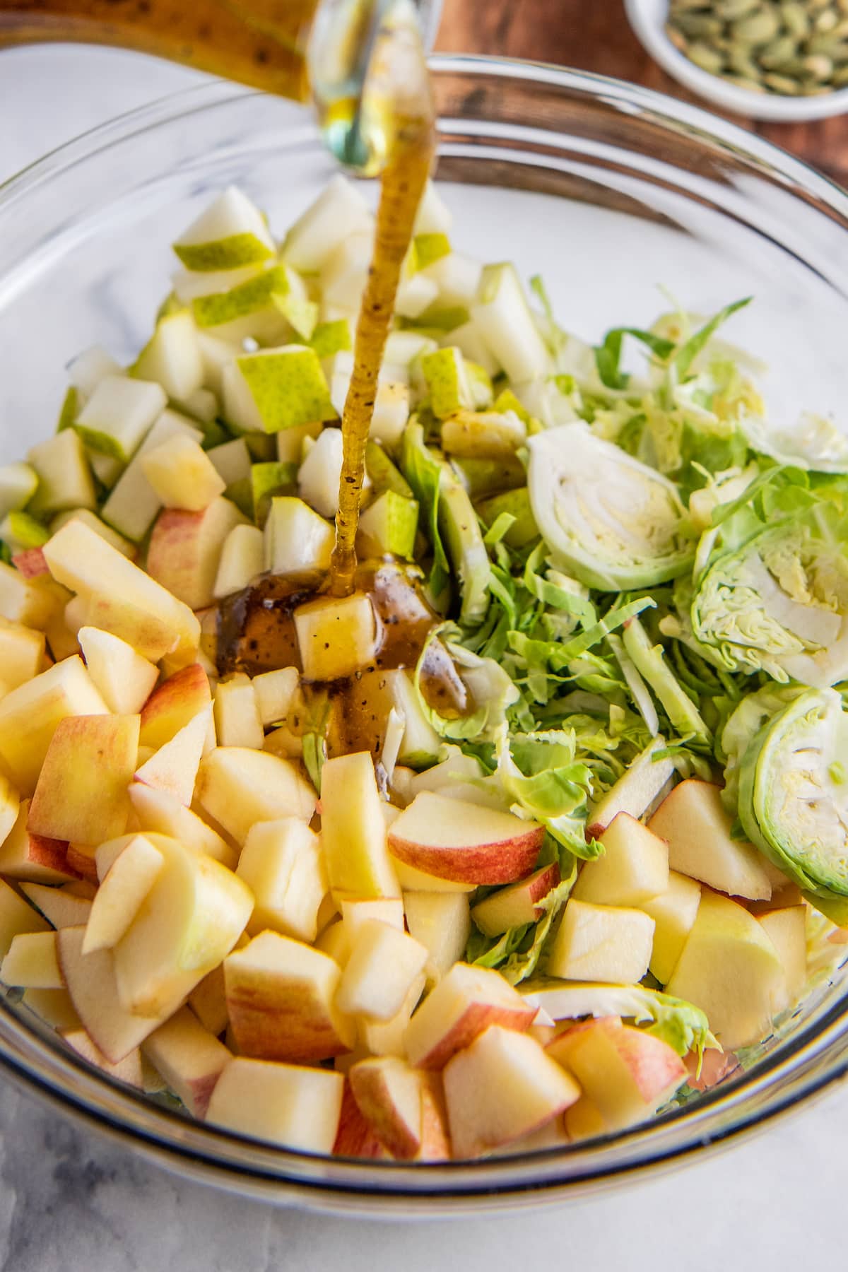 A glass bowl filled with apples and shaved Brussel sprouts with salad dressing being poured on top.