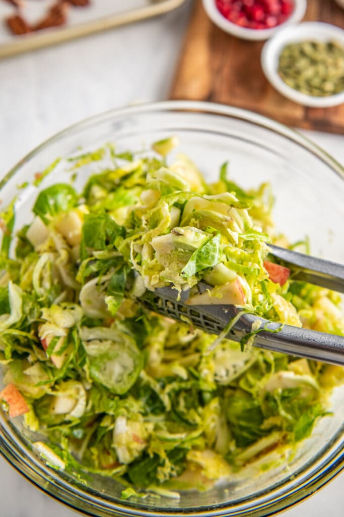 A bowl filled with Shaved Brussel Sprout Salad with tongs tossing the salad.