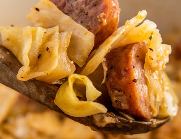 A wooden spoon is scooping out fried cabbage and sausage out of a skillet.