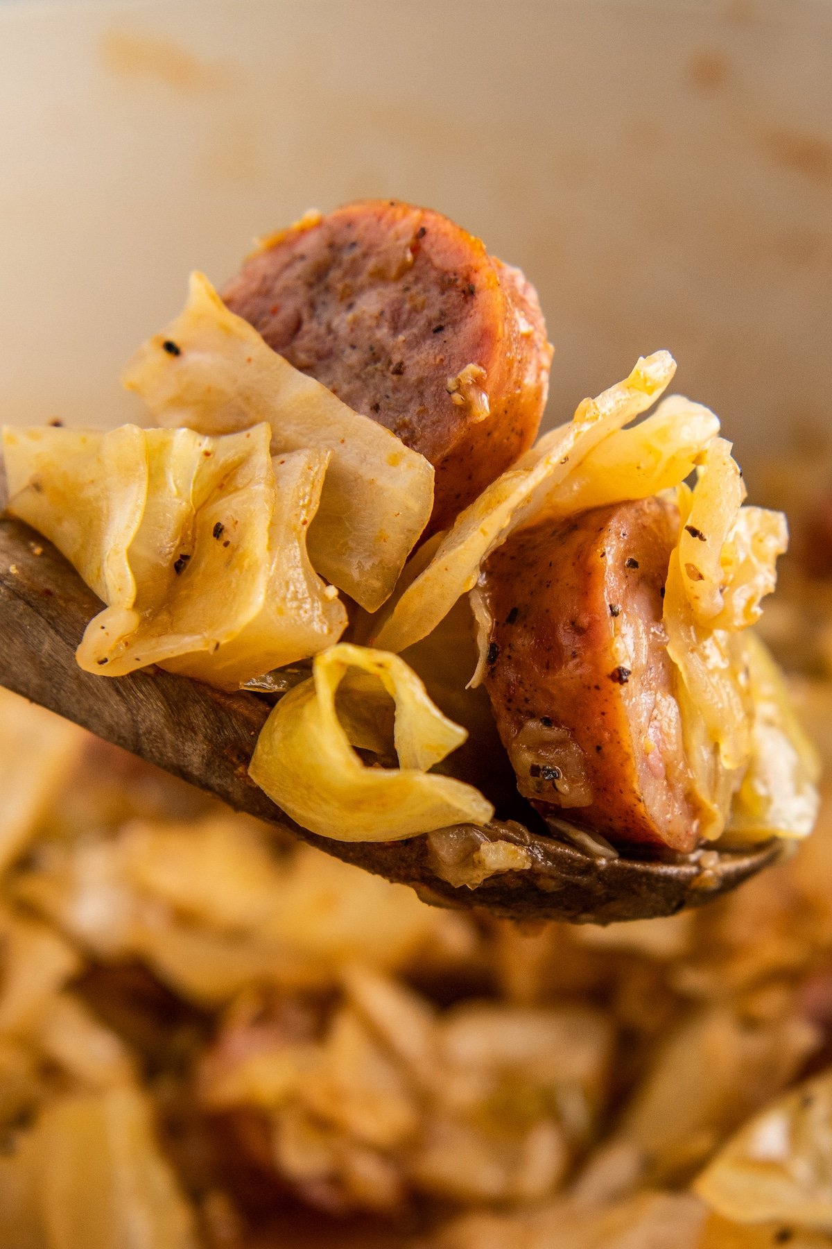A wooden spoon is scooping out fried cabbage and sausage out of a skillet.