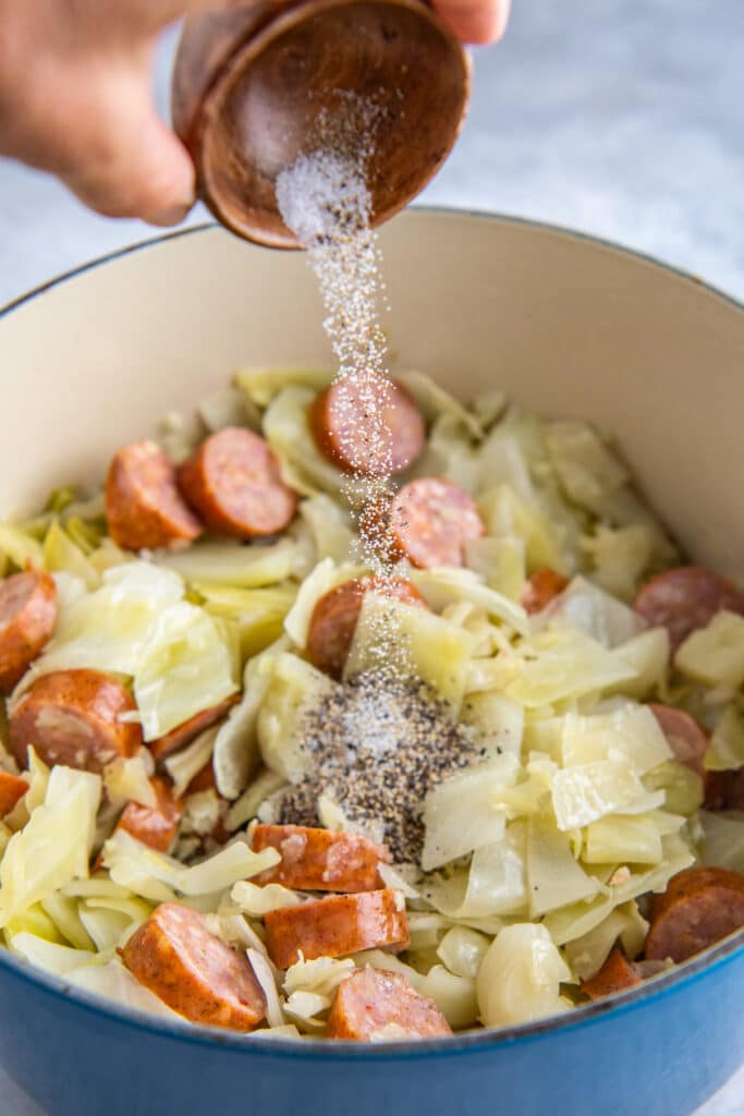 Salt and pepper being poured into a dutch oven filled with fried cabbage and sausage.