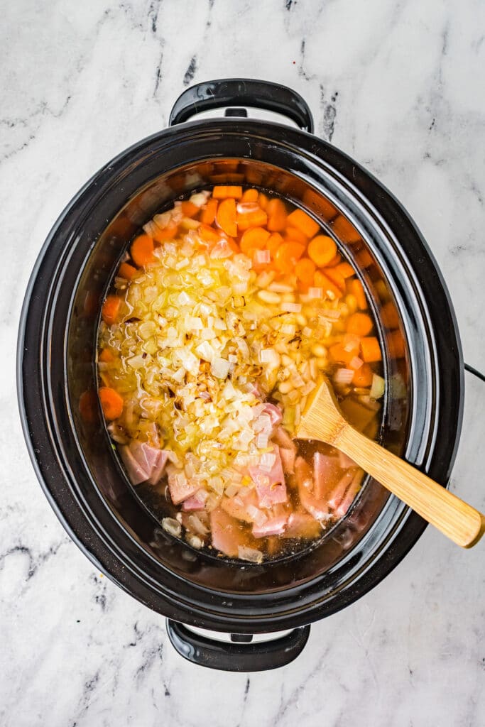 Ham, onions, carrots, and broth added to a crockpot bowl with a wooden spoon.