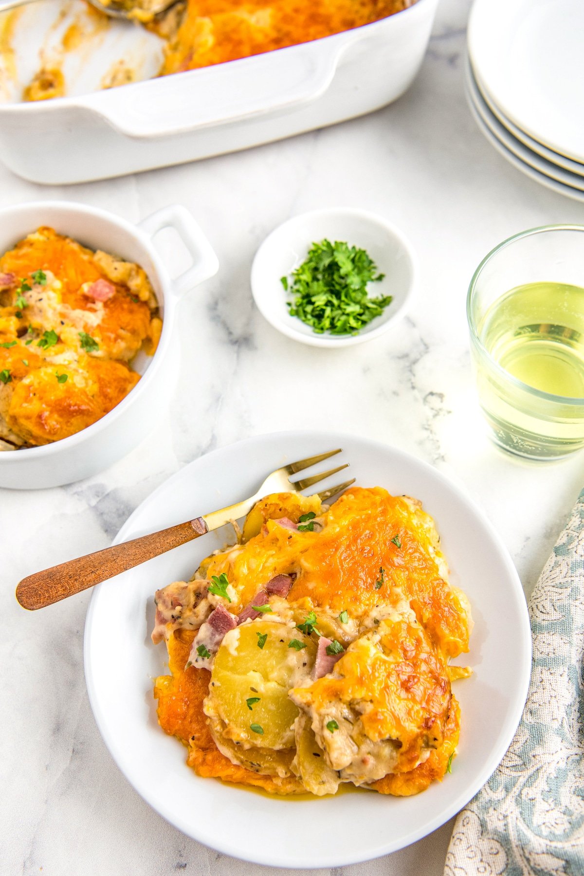 A plate filled with scalloped potatoes with ham casserole on a white plate with a napkin and a glass of wine.