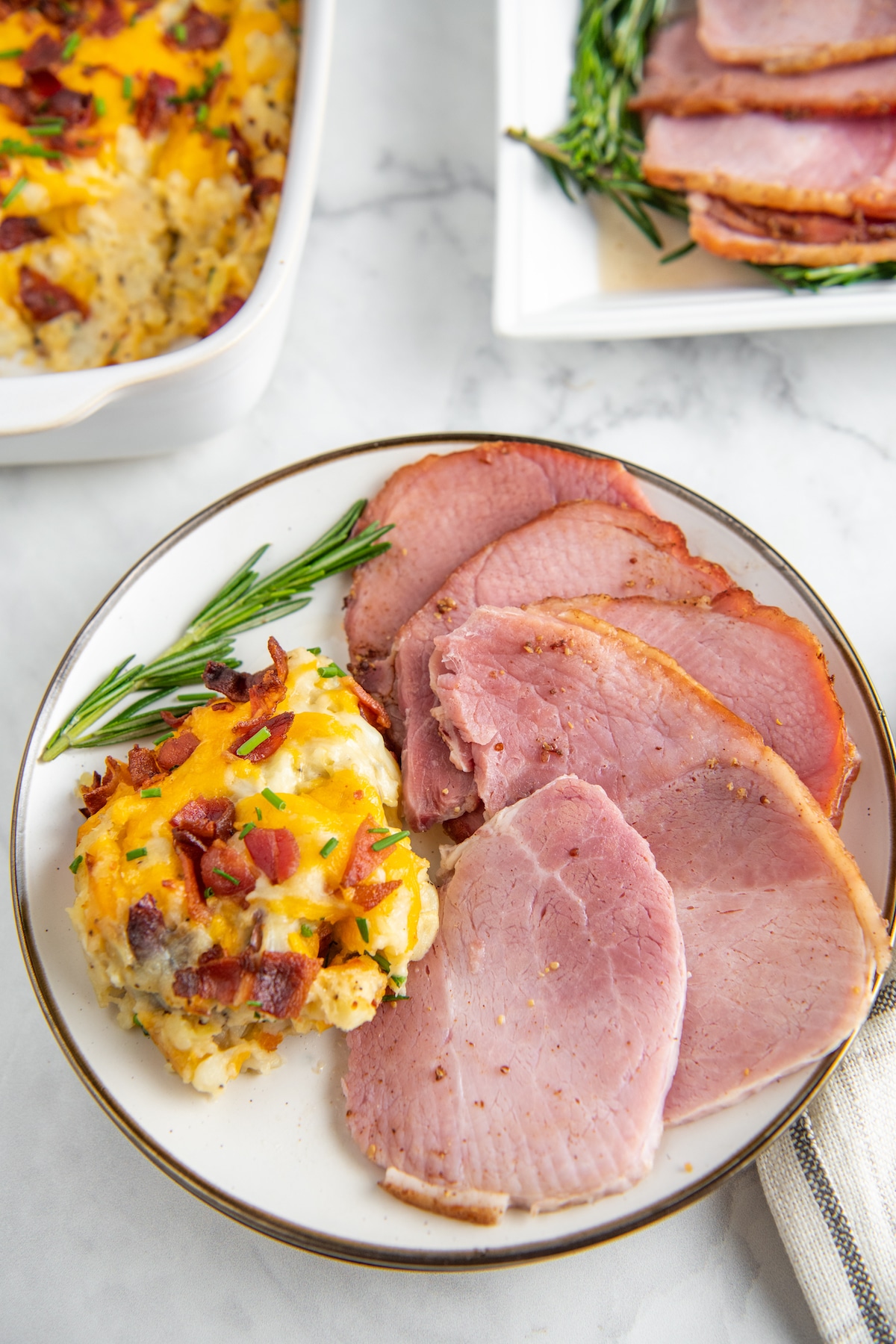 Slices of ham on a white plate with mashed potatoes and fresh rosemary.
