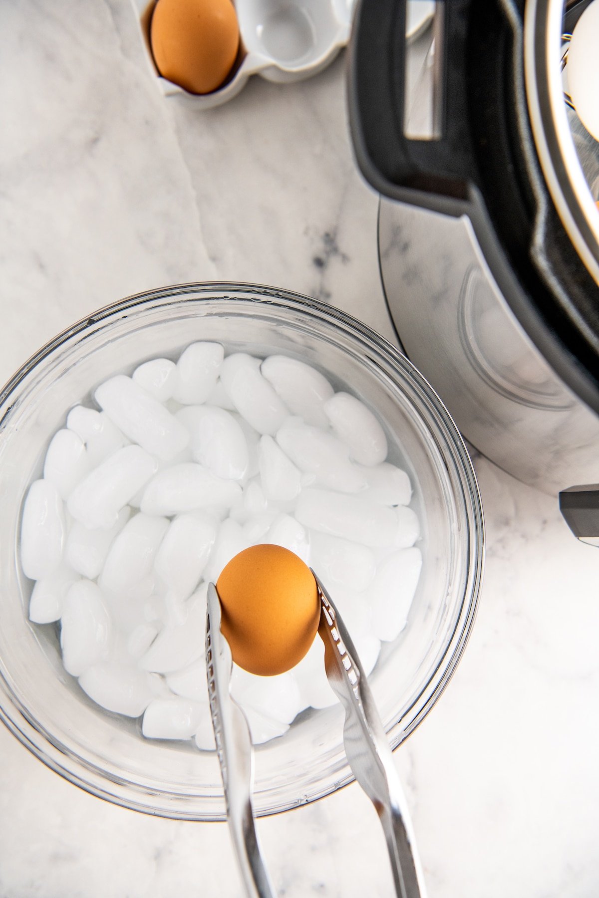 A bowl of ice water with a hard boiled egg being added to it with tongs.