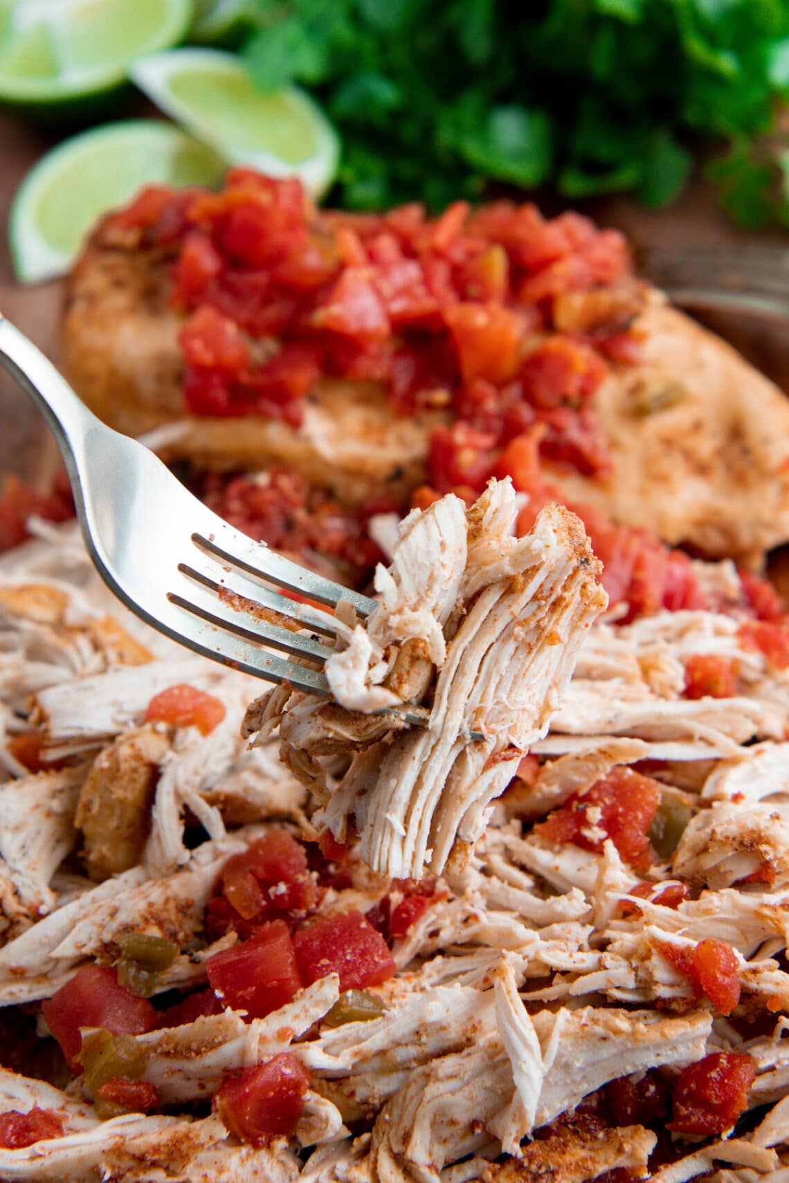 Shredded chicken with tomatoes on a cutting board with a fork picking up a piece of shredded chicken.