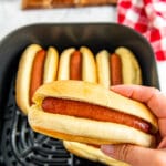 A hot dog in a bun being held with an air fryer with hot dogs in it.