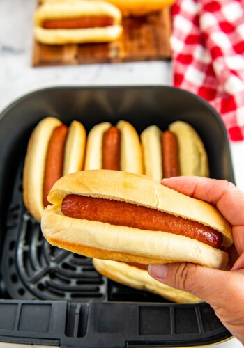 A hot dog in a bun being held with an air fryer with hot dogs in it.
