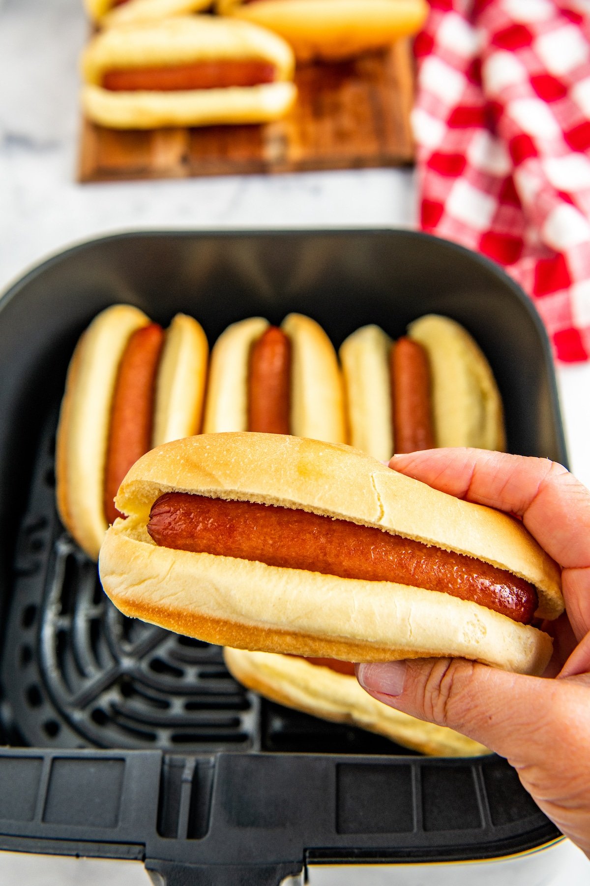 A hot dog in a bun being held with an air fryer with hot dogs in it.