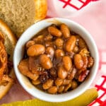 Baked beans in a small bowl in a red basket with a pickle and a sandwich.