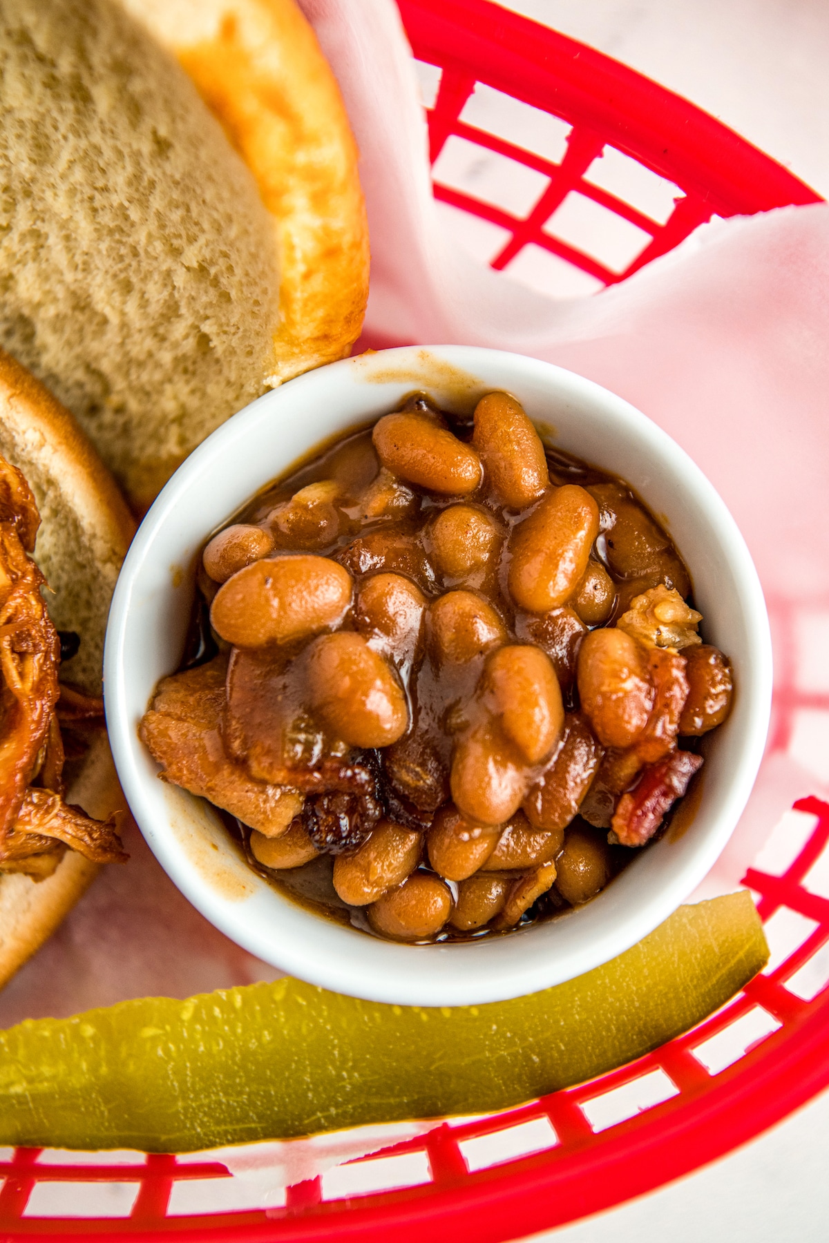 Baked beans in a small bowl in a red basket with a pickle and a sandwich.