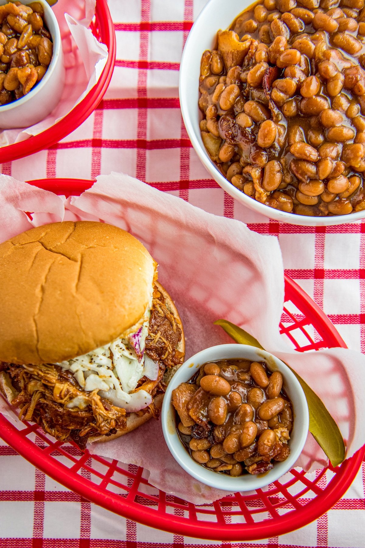 Baked beans in bowls with a pulled pork sandwich and pickle.