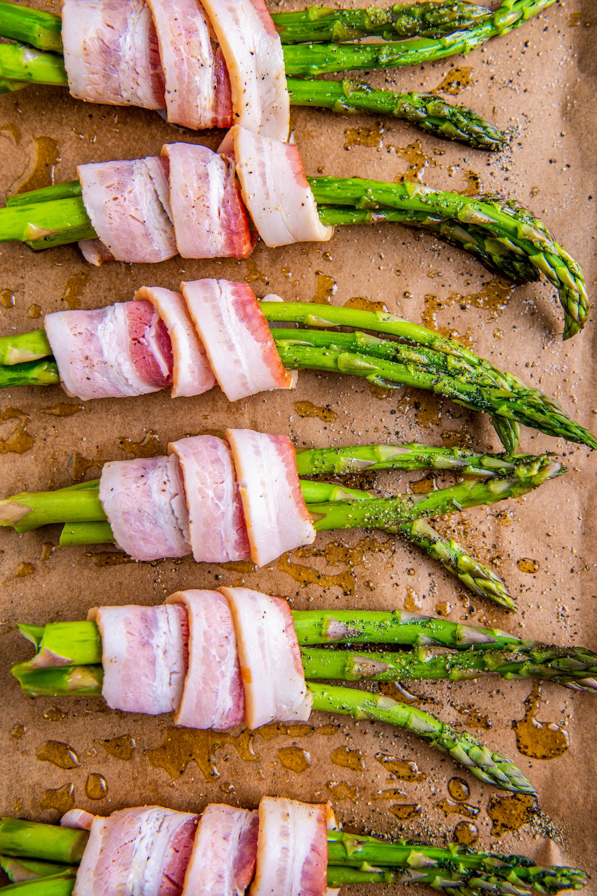 Asparagus wrapped with bacon before baking on a cookie sheet with olive oil being drizzled on top.