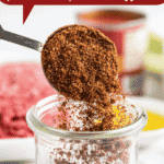 Chili seasoning in a glass jar with a measuring spoon scooping up some of the seasoning mix.