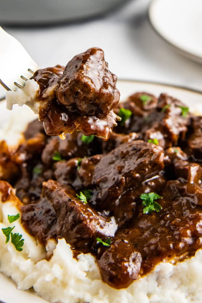 A fork lifting a bite of beef tips and beef tips on a bed of mashed potatoes.