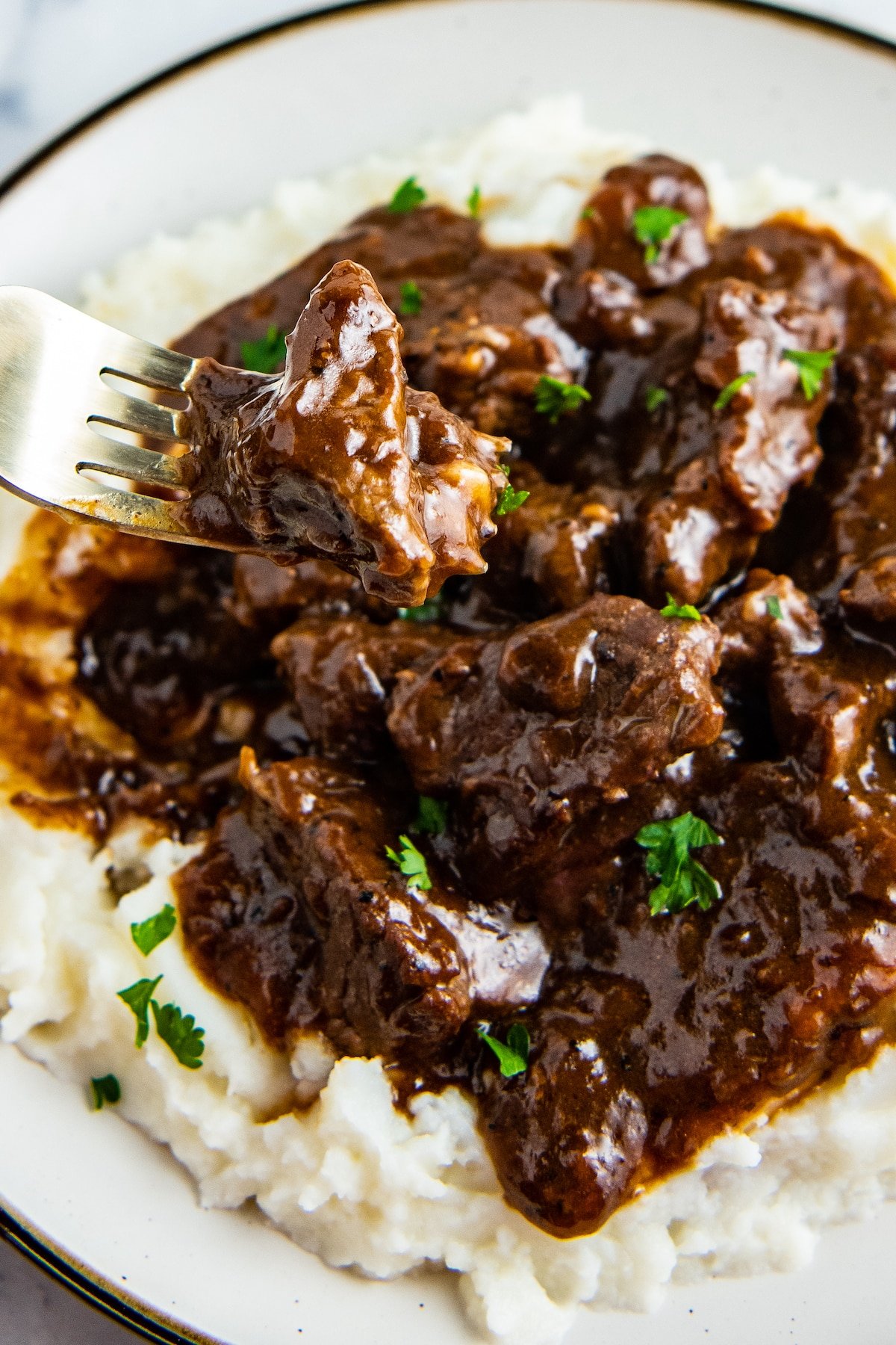 A fork lifting a bite of beef from a plate of beef tips in gravy on mashed potatoes.