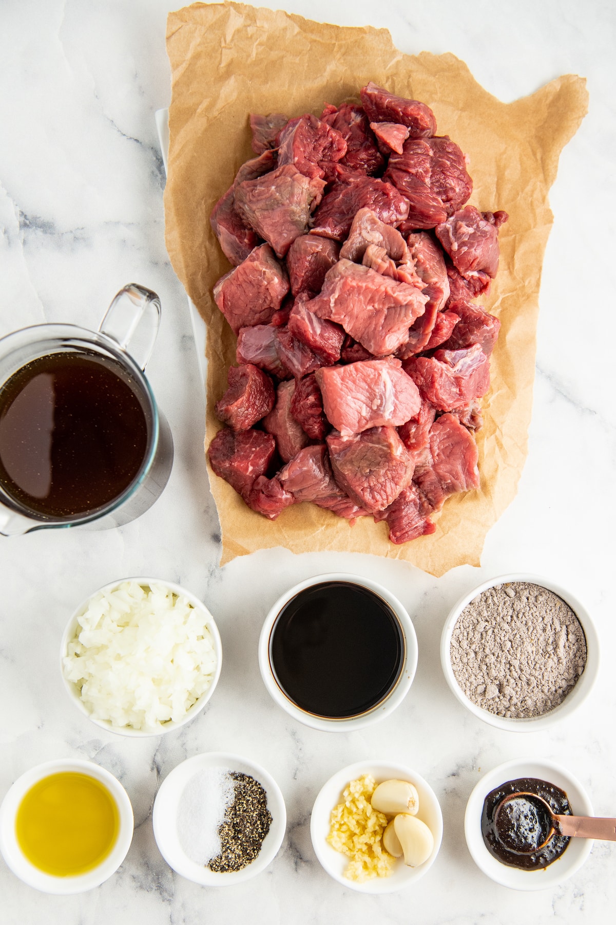 Ingredients in white bowls and beef cut into pieces on parchment paper.