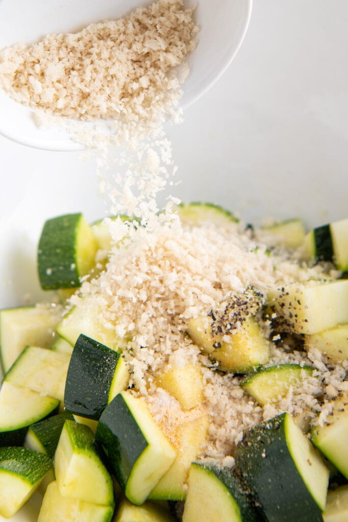 Zucchini in a bowl with salt and pepper and breadcrumbs being poured on top.