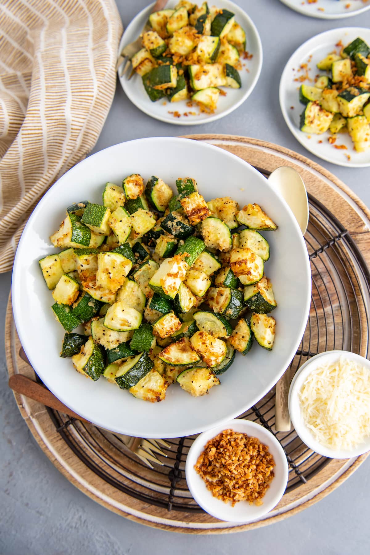 A large bowl of air fryer zucchini with two plates of zucchini and small bowls filled with parmesan cheese and crispy bread crumbs.