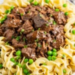 A plate with egg noodles and beef tips on top with peas and parsley on top.
