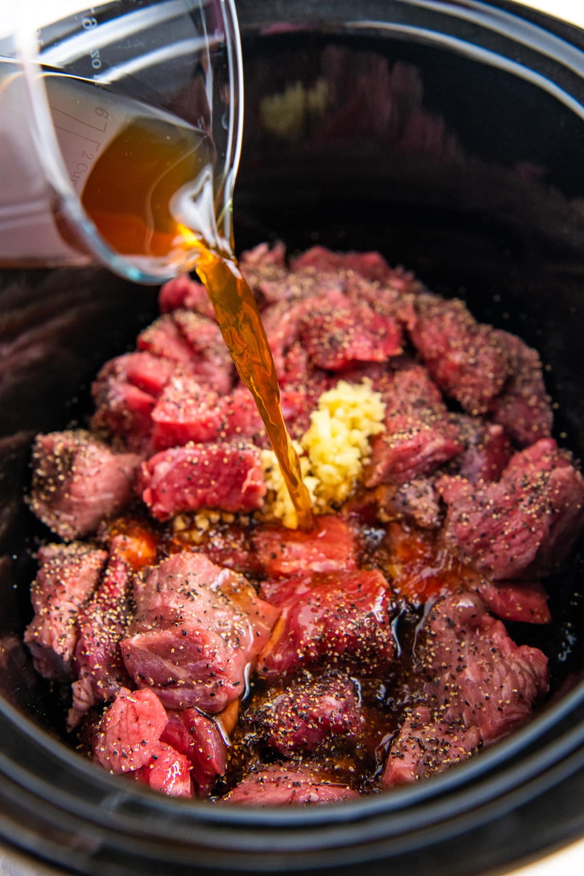 Beef broth being poured into a crockpot with beef tips inside.