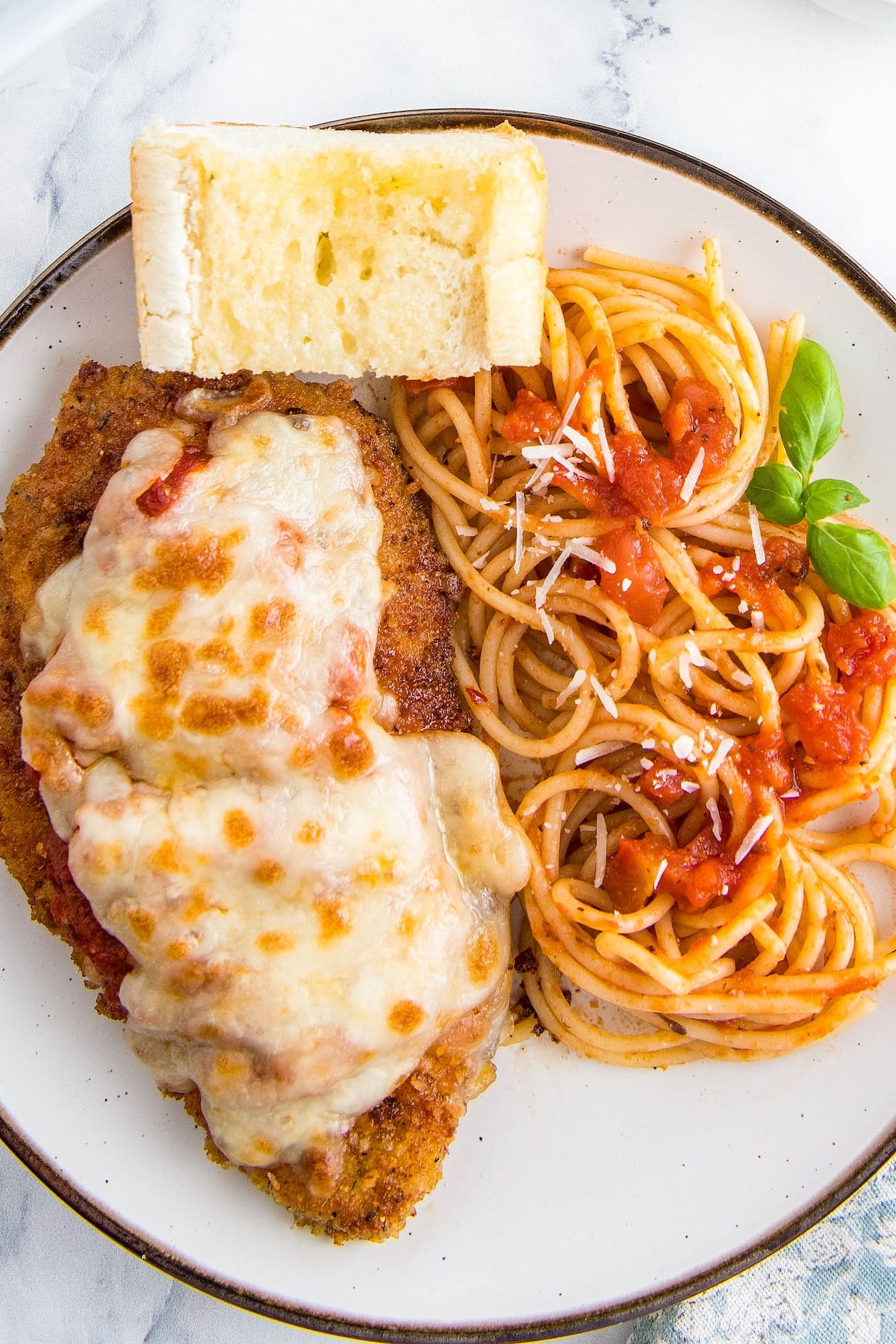 Chicken parm, spaghetti and a piece of garlic bread on a white plate.