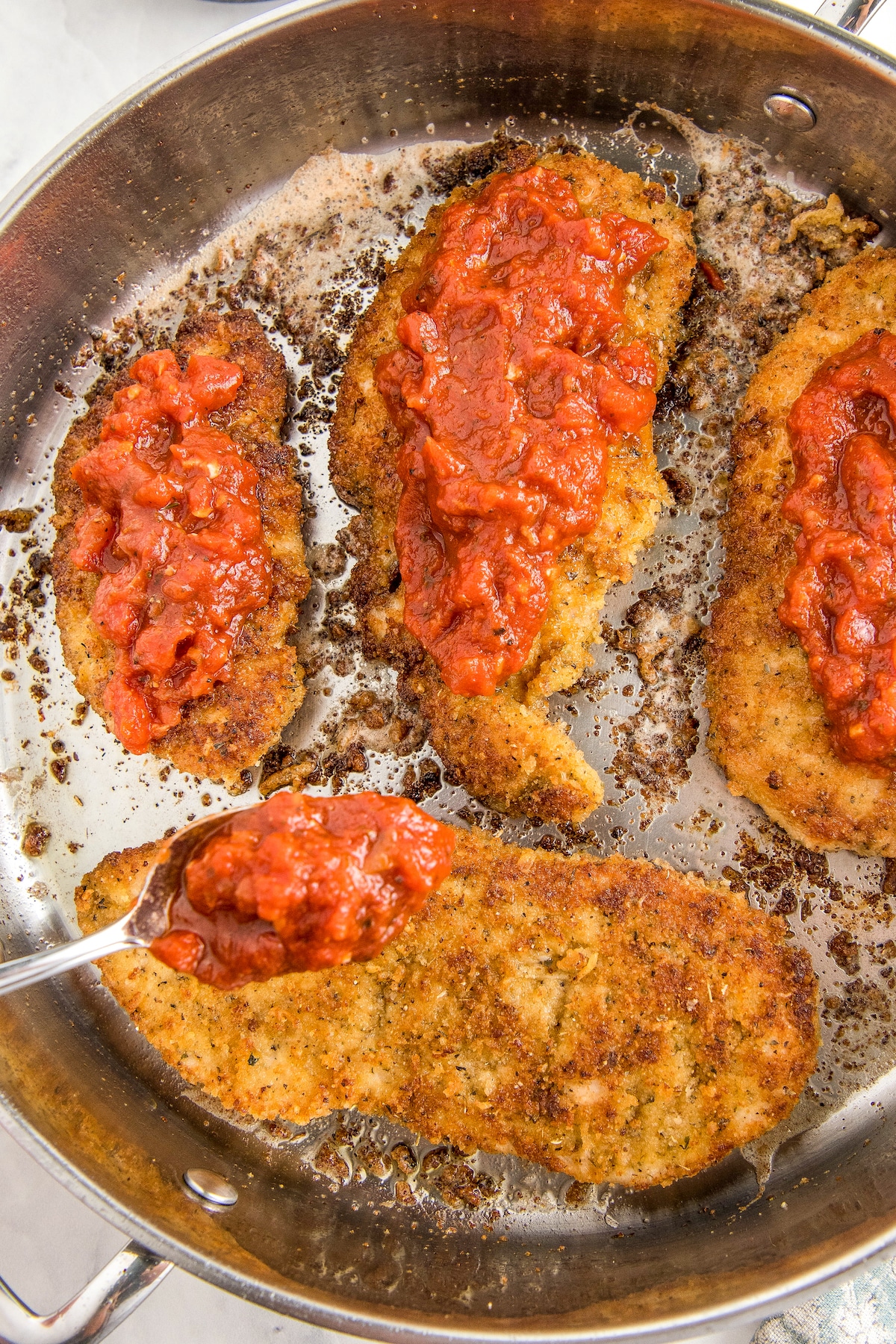 Pan fried chicken breasts being topped with marinara sauce.