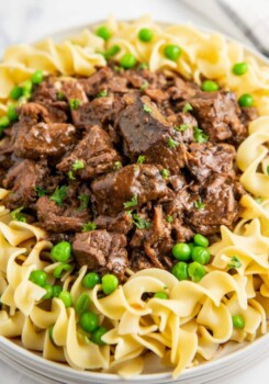 A plate with egg noodles and beef tips on top with peas and parsley on top.