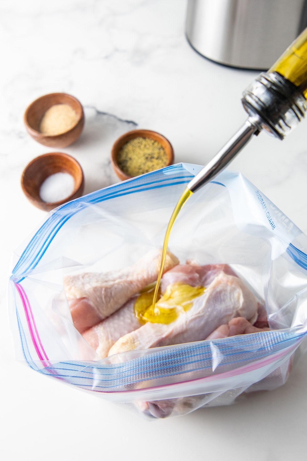 pouring oil into a plastic bag with chicken drumsticks and three prep bowls with seasonings on the side