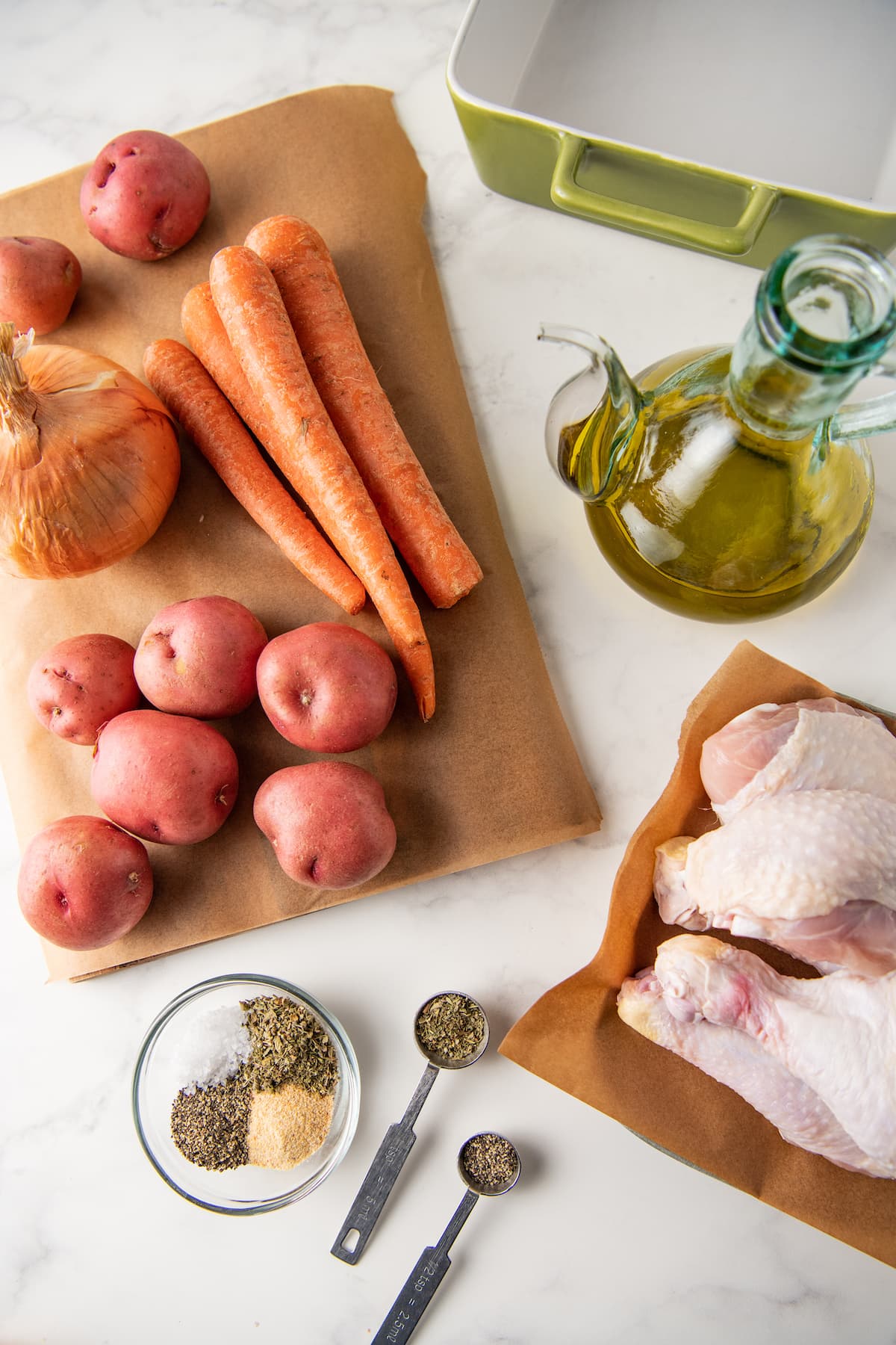 prep set up with a cutting board with potatoes, carrots, and onions, plus chicken legs, seasoning, and olive oil