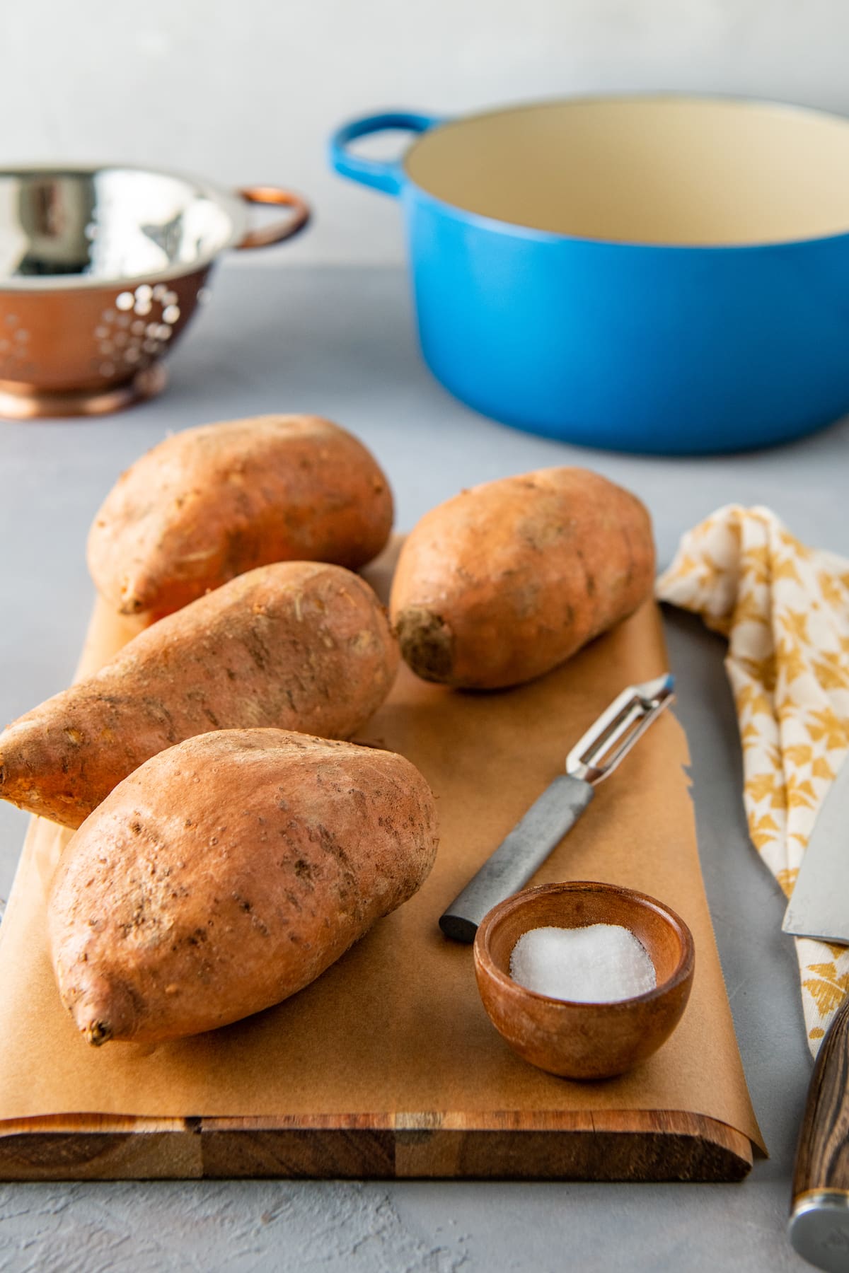 whole sweet potatoes on a cutting board with salt and a peeler