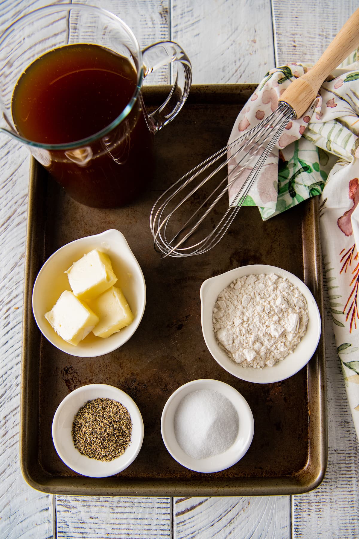 ingredients to make homemade brown gravy on a sheet tray in small prep bowls including butter, flour, salt, and pepper. plus a measuring cup with beef stock.