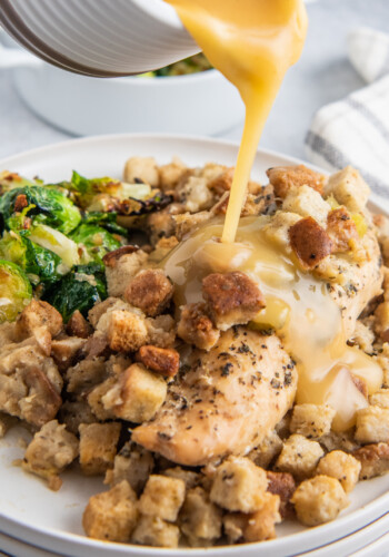 gravy being poured over a plate of cooked chicken with stuffing and a side of Brussels sprouts
