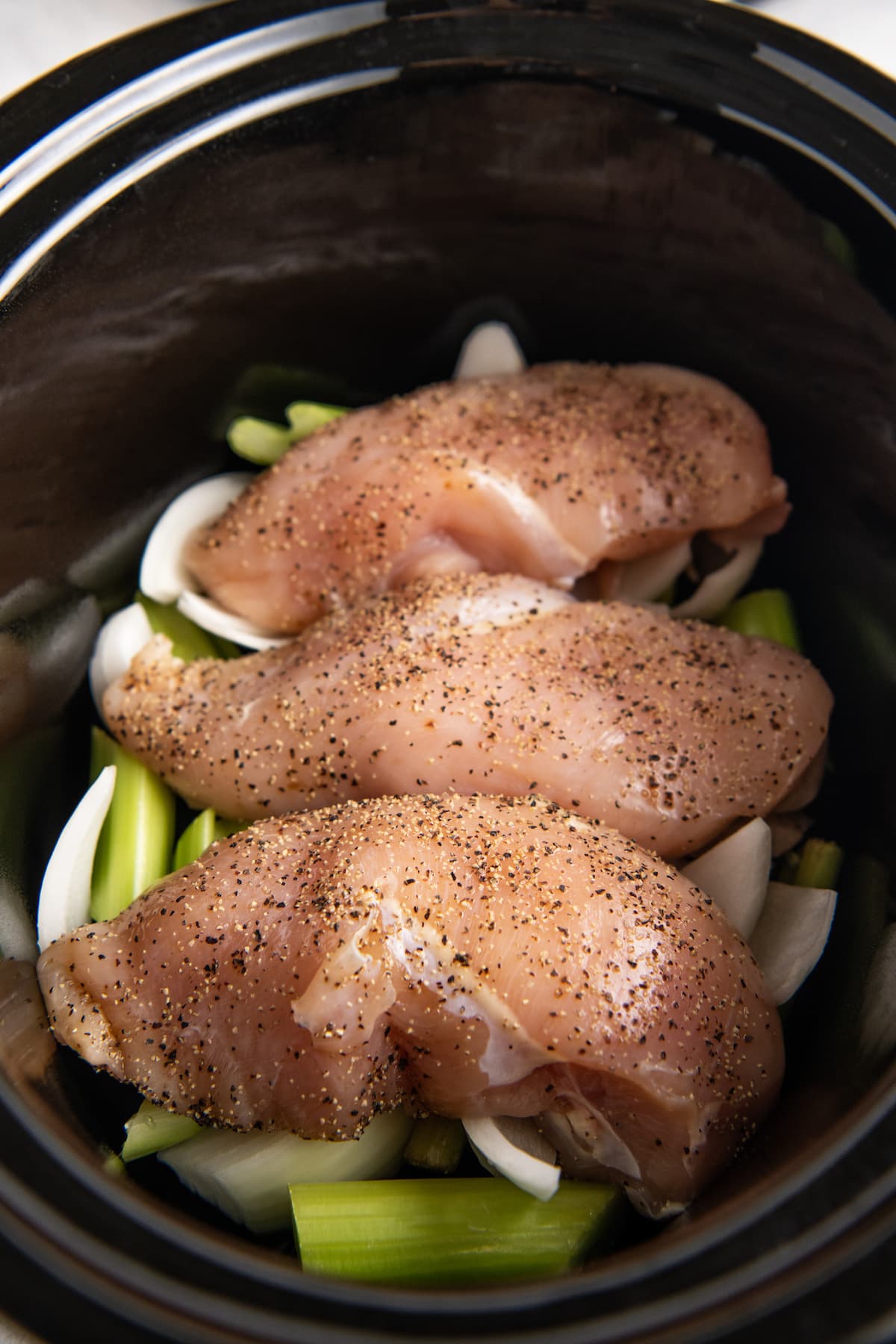 three seasoned chicken breasts in a black pot on top of chopped celery and onions