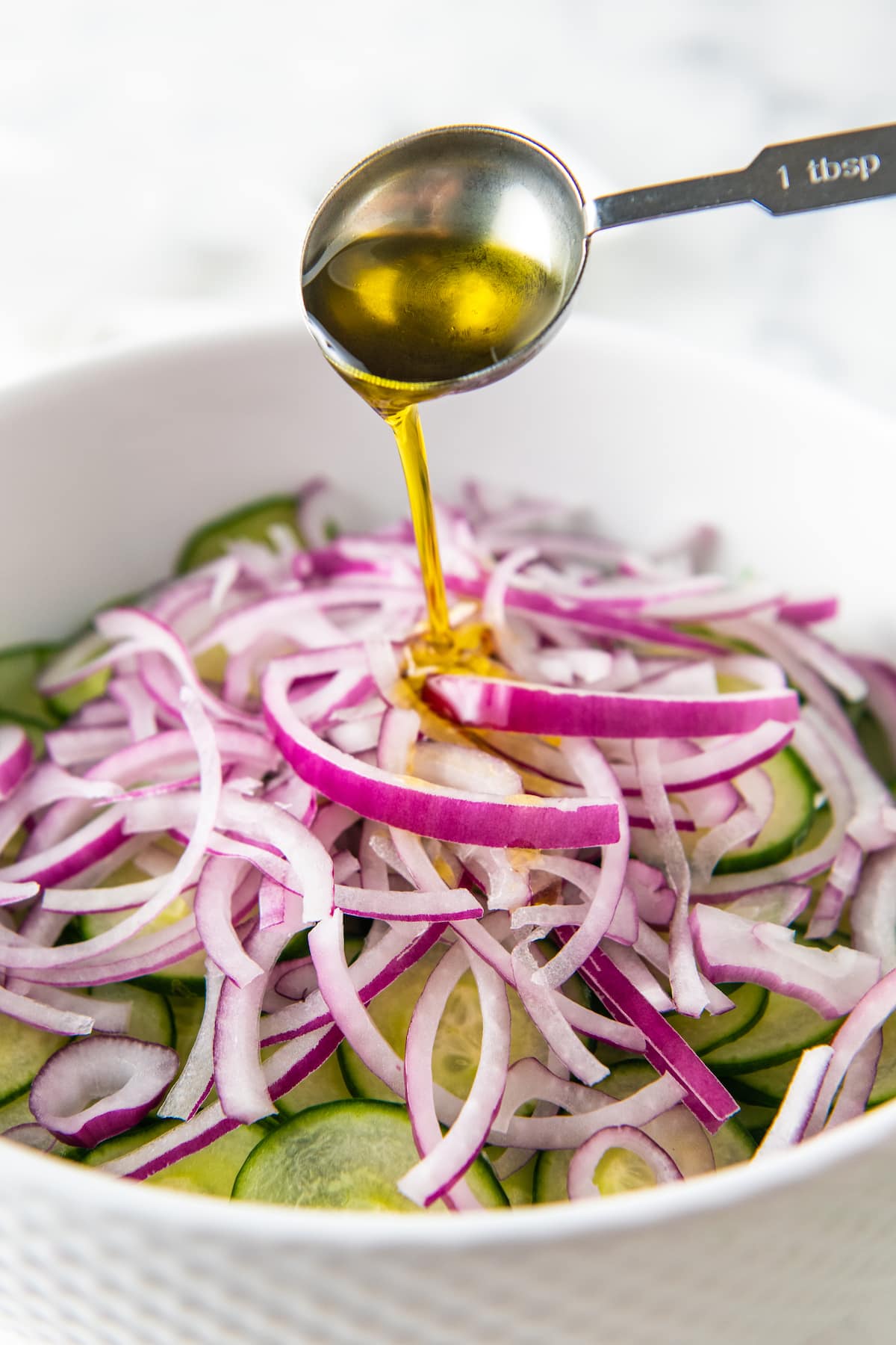 Olive oil being poured over a bowl of sliced cucumbers and red onions.