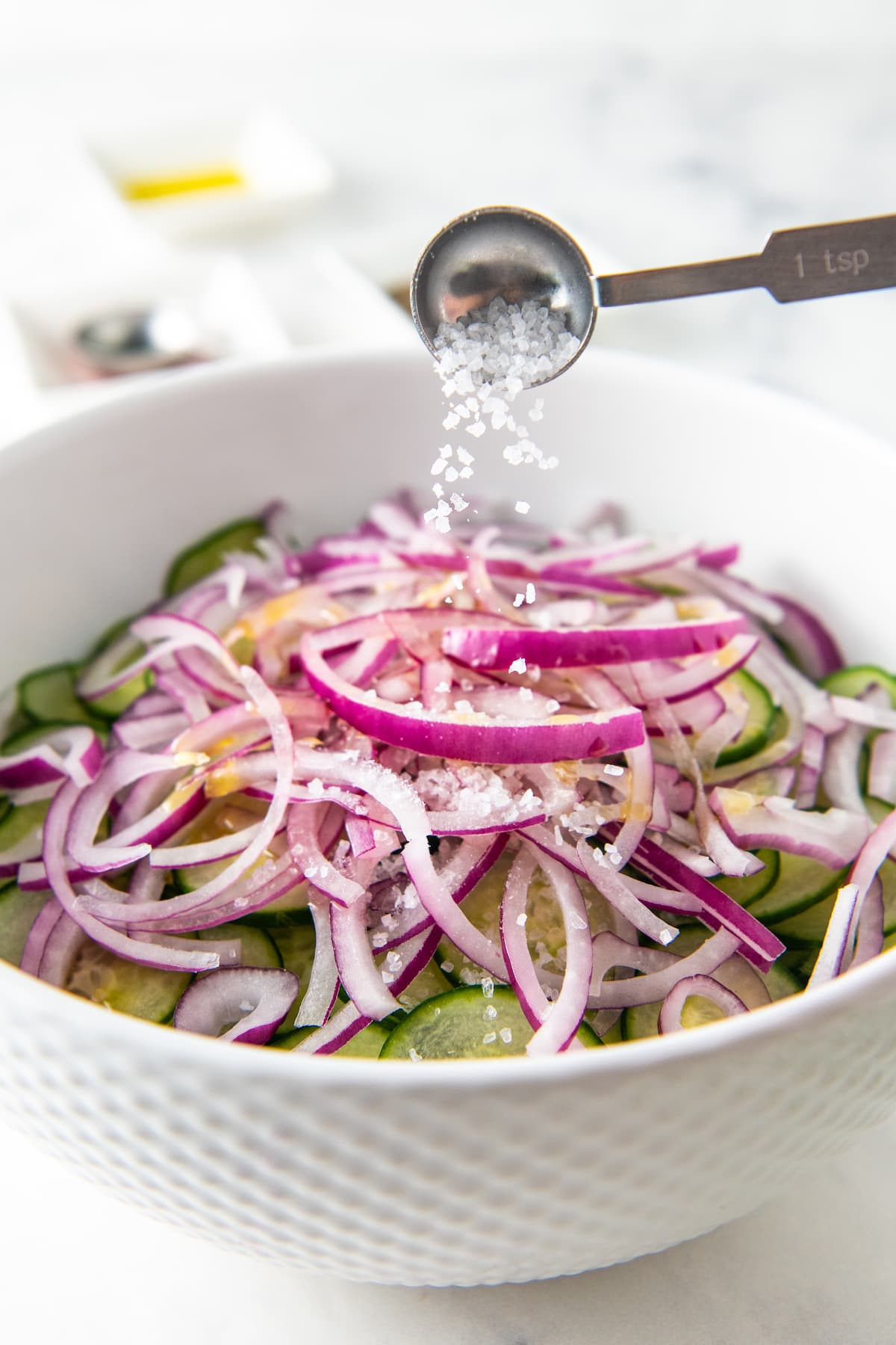 Salt being added to a bowl filled with cucumbers, red onions and olive oil.
