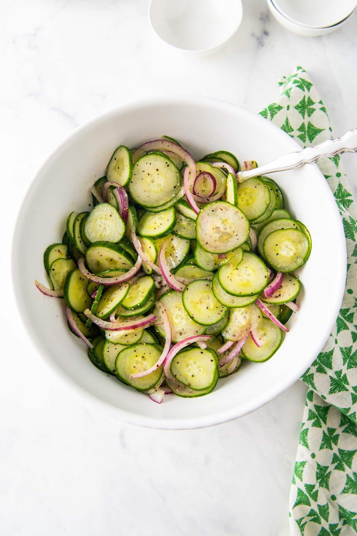A big white bowl filled with cucumber salad with red onion with a spoon and tea towel.