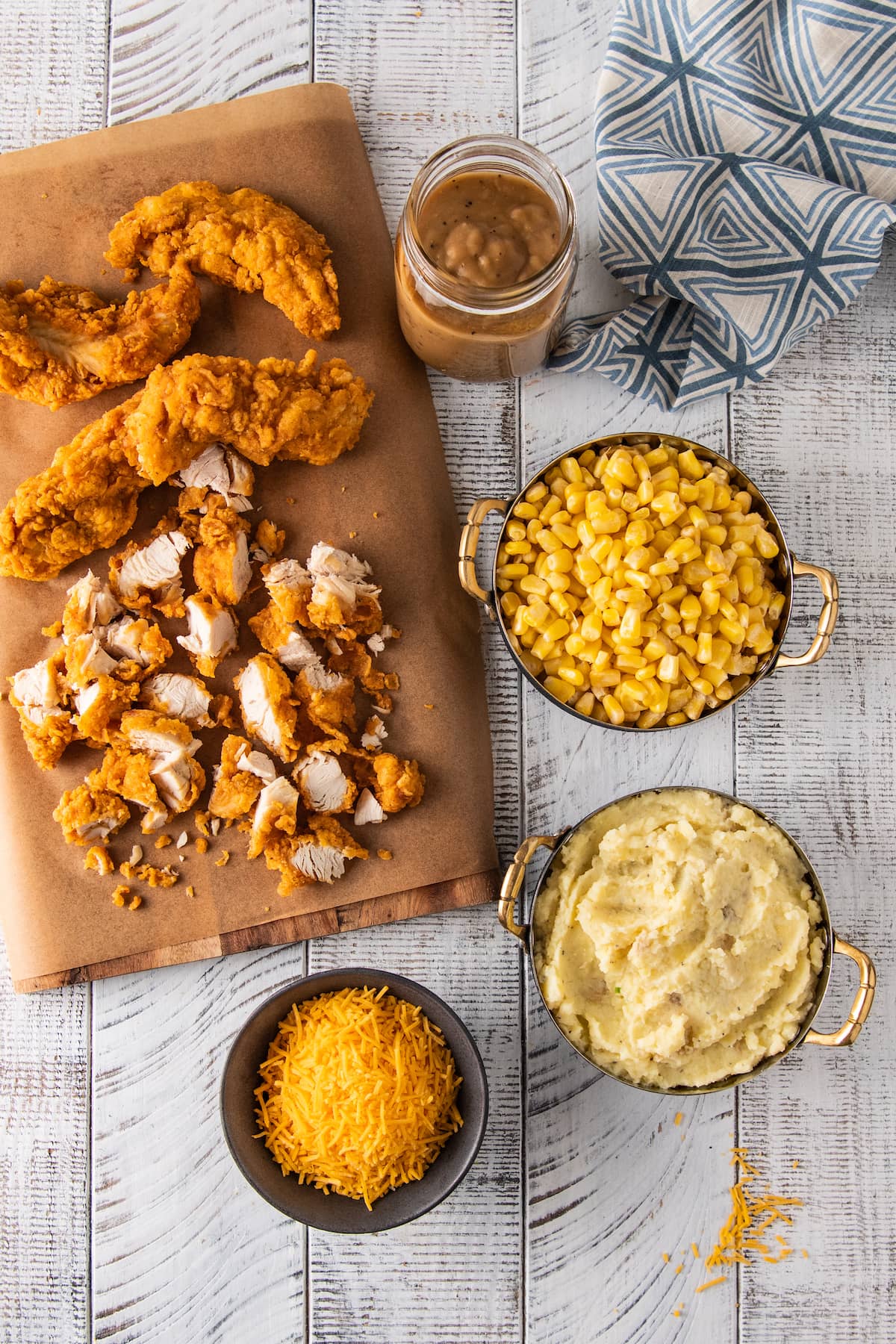 the ingredients to make a fried chicken casserole dish based off of the KFC Bowl, there's corn, mashed potatoes, cheese, gravy, and fried chicken chopped on a cutting board