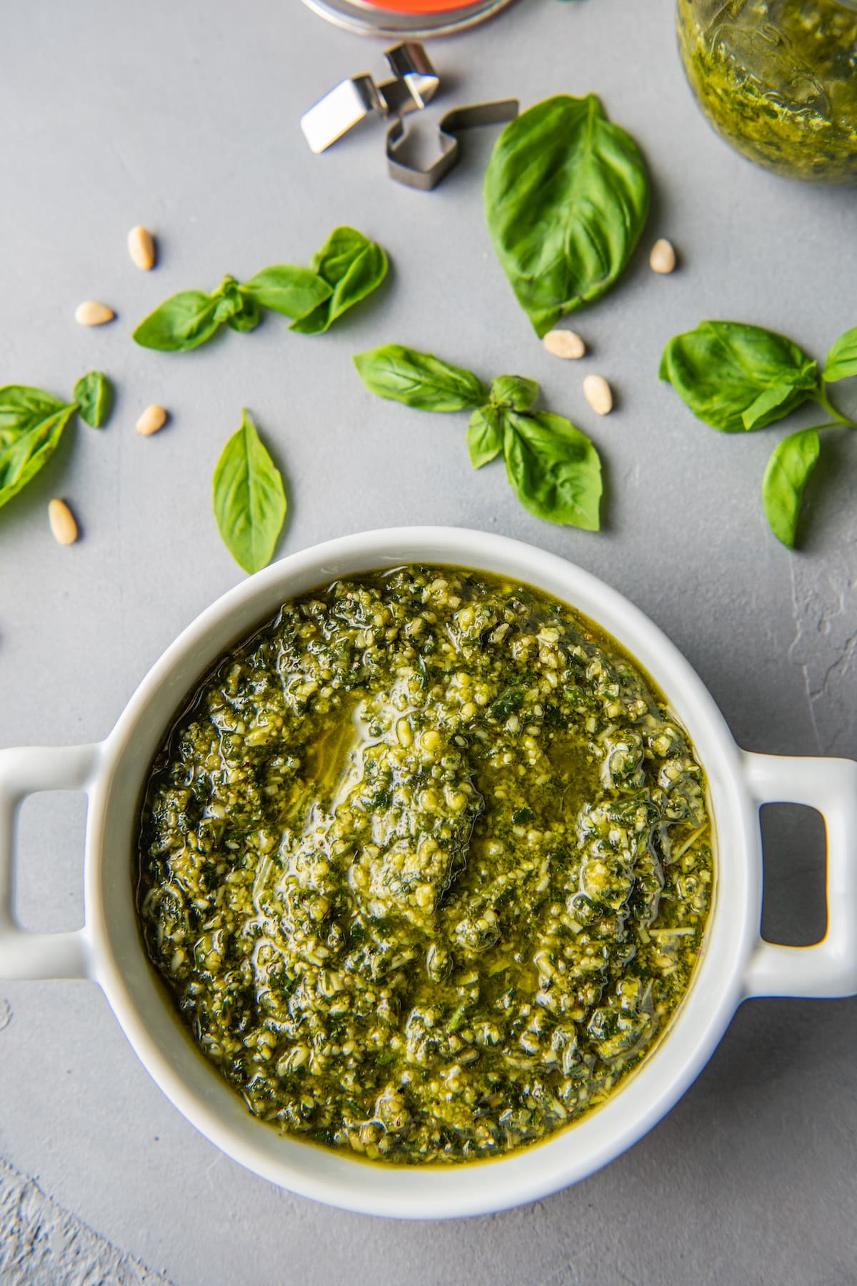 fresh basil pesto in a white bowl with basil leaves and pine nuts scattered next to it