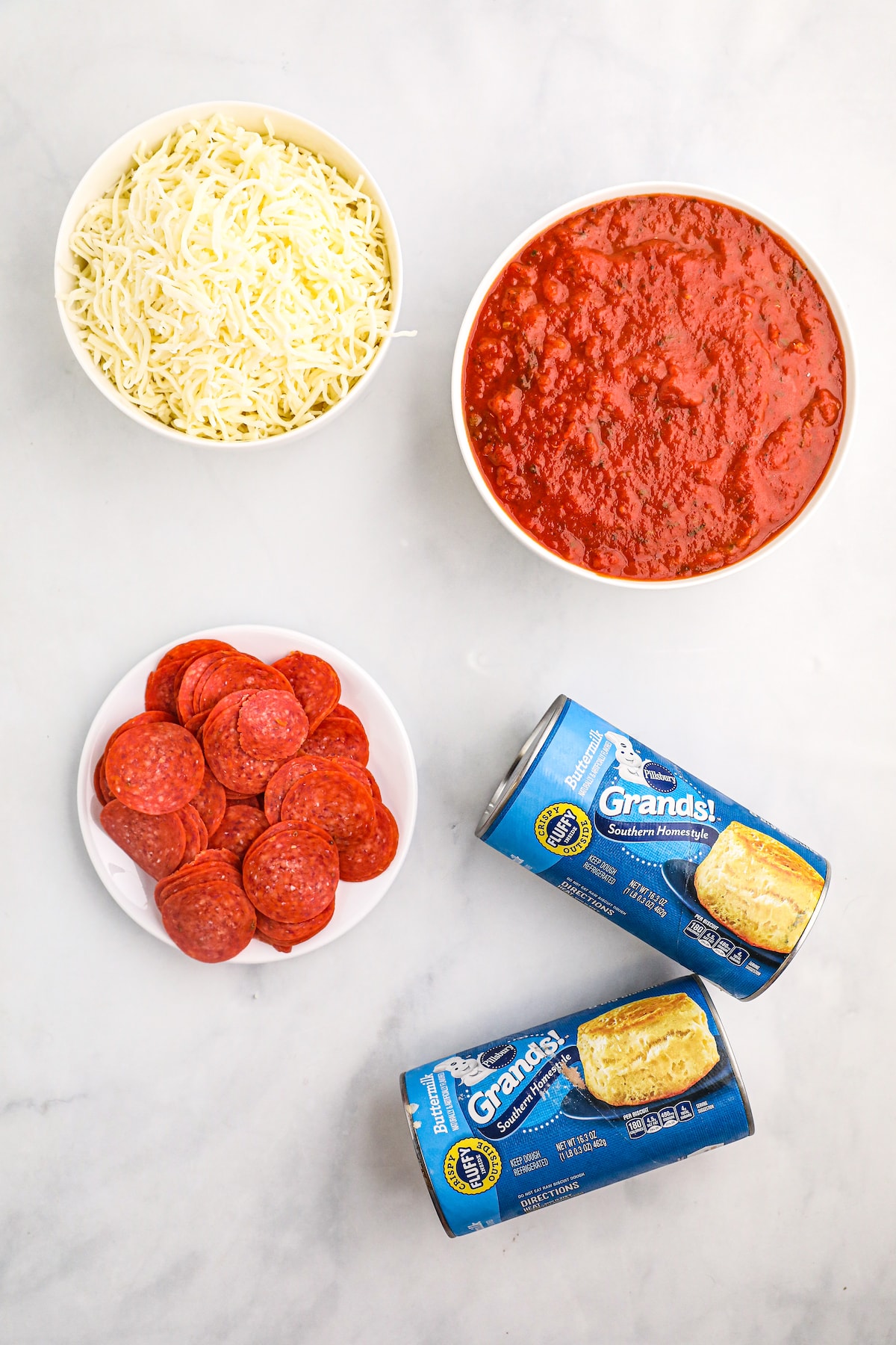 three bowls with shredded cheese, tomato sauce, and pepperoni next to two containers of refrigerated biscuit dough